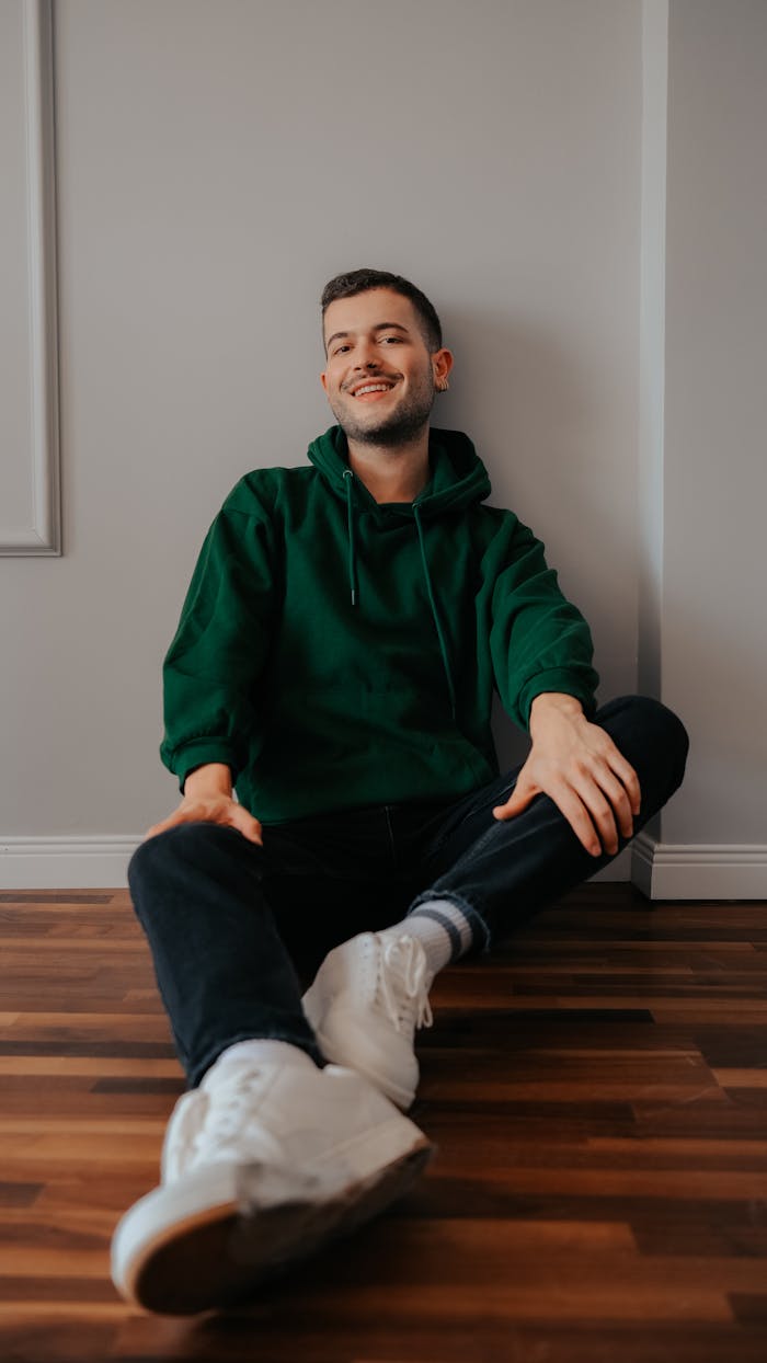 about-img-01 Smiling young man in a green hoodie sitting on a wooden floor indoors.
